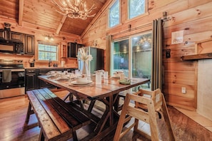 A wooden cabin interior with a dining table set for four, a kitchen with modern appliances, and a large window.