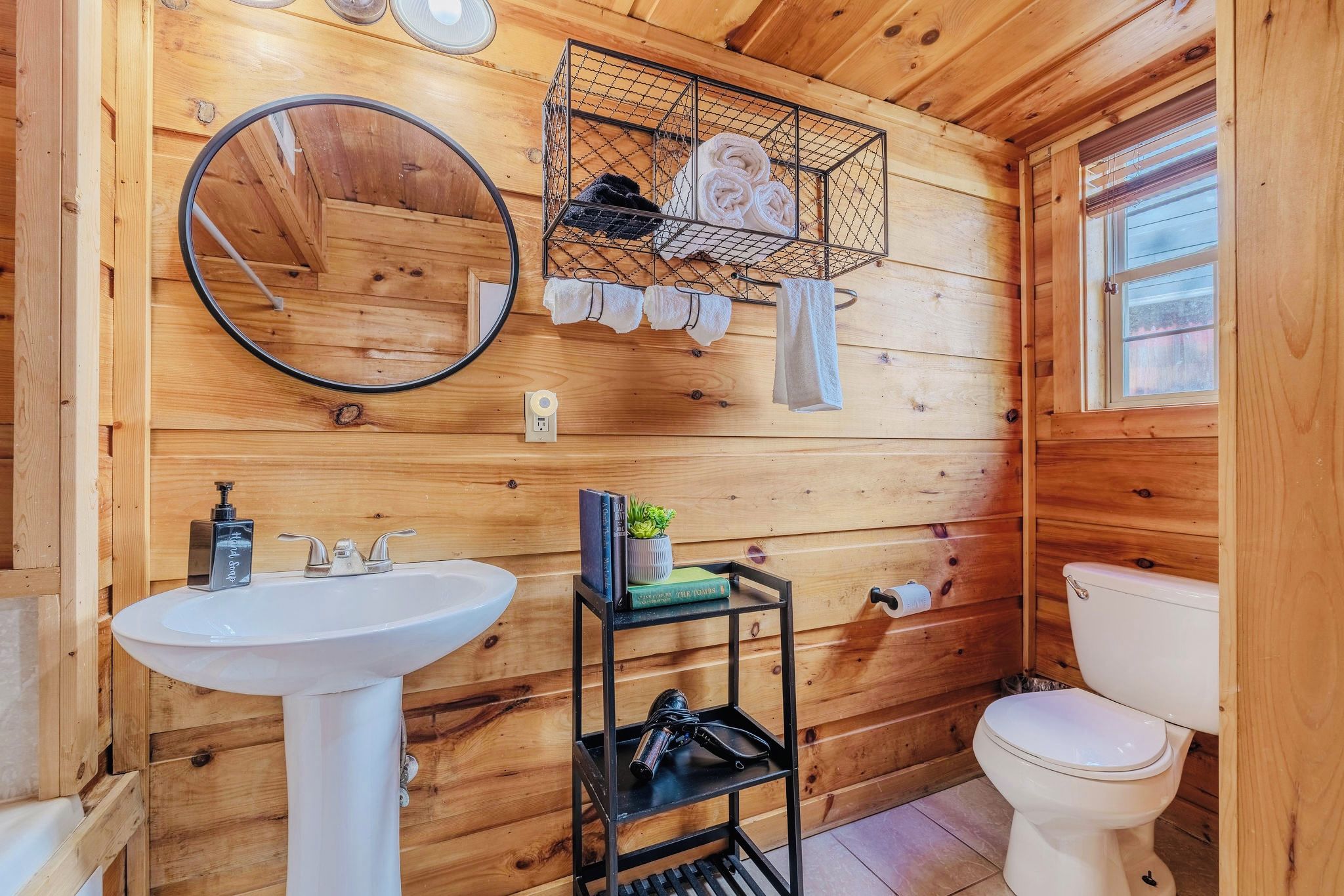 A wooden cabin bathroom with a white sink, a toilet, a mirror, and a shelf with towels and toiletries.