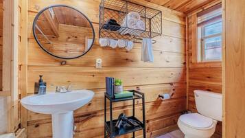 A wooden cabin bathroom with a white sink, a toilet, a mirror, and a shelf with towels and toiletries.