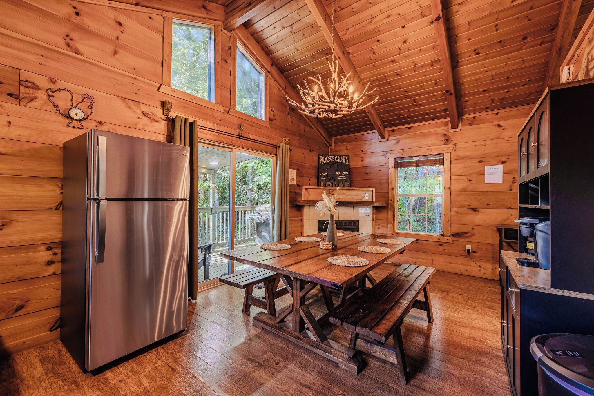 A wooden cabin interior with a stainless steel refrigerator, a wooden dining table with benches, a chandelier, and a fireplace.
