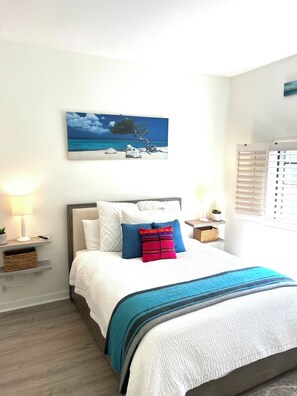 Un letto rifatto con un copriletto bianco e una coperta a righe blu e nere, un quadro raffigurante una spiaggia sulla parete e una finestra con persiane.