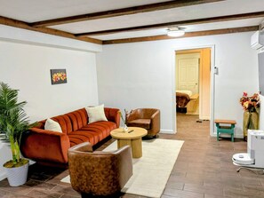Ein Wohnzimmer mit einer Couch, einem Sessel und einem Couchtisch, einer Zimmerpflanze und einer Tür zu einem anderen Raum.