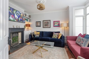 A living room with a dark blue sofa, a glass coffee table, a fireplace, and artwork on the walls. - The Tri-House (Watford)