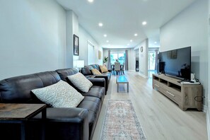 A modern living room with a leather sofa, a coffee table, a TV, and a dining area with chairs and a table. - Spacious 4-Bedroom Home Sleeps 8 Backyard (Barrie)