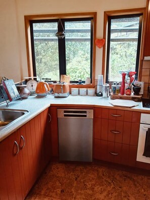 A kitchen with wooden cabinets, a stainless steel dishwasher, and a window with a view of greenery. - Shell House - A One-of-a-Kind Stay (Mangōnui)