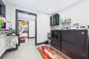 A compact hotel room with a kitchenette, including a washing machine and dryer, a small shelf with toiletries, and a door leading to another room. - Shimmering Peaks Retreat (Thornton)
