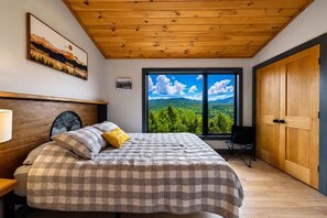 A bedroom with a large window, a bed with a checkered bedspread, a wooden ceiling, and a door. - Shimmering Peaks Retreat (Thornton)
