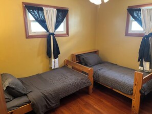 Une pièce avec deux lits superposés en bois, chacun doté d’un matelas et de coussins matelassés gris. Il y a deux fenêtres avec des rideaux attachés, et une tête de lit en bois sur chaque lit.