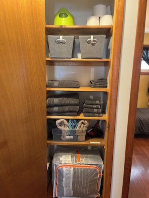 A wooden shelf unit with storage bins, towels, and a plastic container. - Family Home with Large Kitchen & Sun Porch (Erie)
