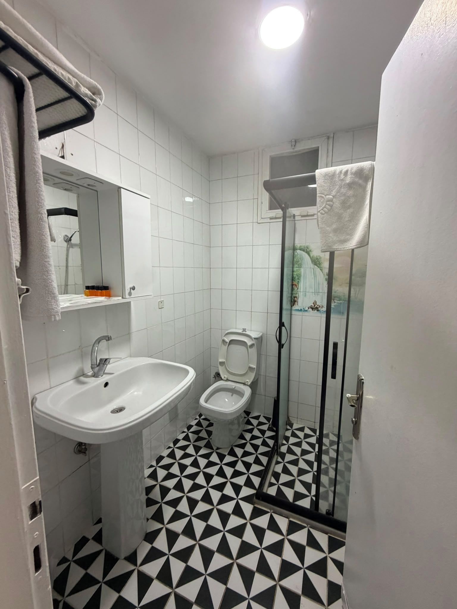 Un baño con lavamanos, inodoro y zona de ducha, con piso de baldosas geométricas blanco y negro.