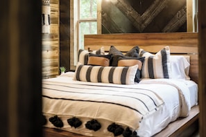A bed with striped bedding and multiple pillows, a wooden headboard, and a window with a view of trees. - Hot Tub | Game Room | Playset | Patio Oasis! (Broken Bow)