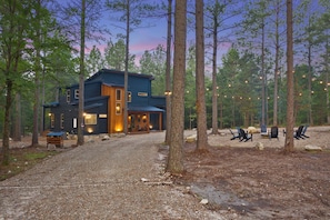 A cabin with a gravel driveway, surrounded by tall trees and outdoor seating. - Hot Tub | Game Room | Playset | Patio Oasis! (Broken Bow)