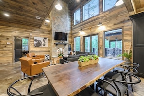A spacious room with a large wooden table, leather chairs, a sofa, and a television mounted on a stone wall. - Hot Tub | Game Room | Playset | Patio Oasis! (Broken Bow)