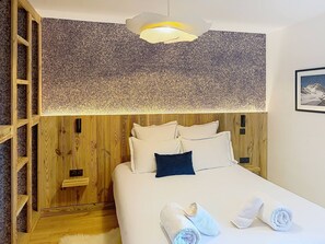 A hotel room with a wooden headboard, a large bed with white bedding, a dark blue pillow, and a wall-mounted light fixture. - Apartment with parking - 4 guests (Montvalezan-La Rosière)