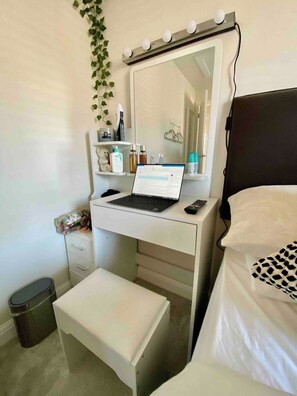 Ein Schlafzimmer mit einem weißen Schminktisch, einem Laptop darauf, einem Bett mit einer gemusterten Decke und einer Mülleimer.