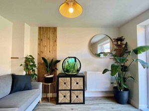 A living room with a sofa, a round mirror, a potted plant, a small table with a decorative item, and a wooden wall panel. - Modern 3Bed House • Parking • WiFi • Hampton Water (Peterborough)