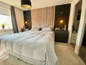 A bedroom with a large bed, bedside tables, a dresser, and a wall-mounted light fixture. - Modern 3Bed House • Parking • WiFi • Hampton Water (Peterborough)