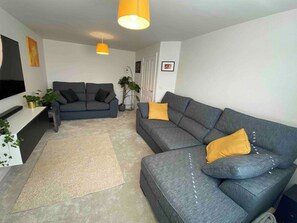 Ein Wohnzimmer mit einem grauen Sofa, einem kleineren grauen Sofa, einem gelben Sitzkissen, einem Fernseher und einer Pflanze.