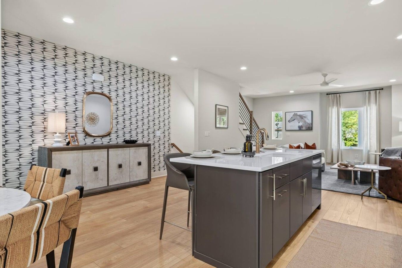 A modern kitchen with a central island, a dining area with chairs, a sofa, and a view of greenery outside.