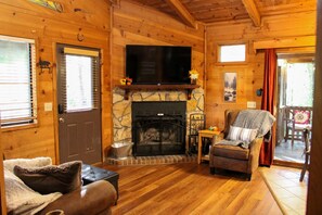 A cozy log cabin interior with a stone fireplace, a television mounted above it, a comfortable seating area with a sofa and armchair, and a door with a window. - 47 Bear Ridge cozy 2 bedroom 1 bathroom cabin. Perfect getaway (Sautee)