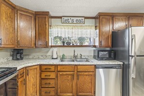 Una cocina con gabinetes de madera, un refrigerador de acero inoxidable, un microondas, una cafetera y una ventana con un cartel que dice 'comamos'.
