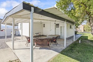 A covered outdoor patio with a table and chairs, a white building in the background, and a grassy area. - Cozy Salina Home w Patio, Yard & Full Kitchen! (Salina)