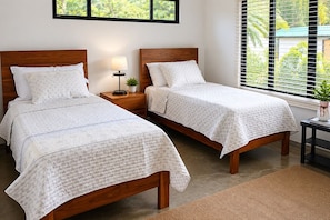 A hotel room with two wooden beds, a nightstand with a lamp, a small table with a plant, and a window with blinds. - Vista Dorada: Spacious 2BR Retreat with Breathtaking Mountain & Sunset Views (San Ramon)