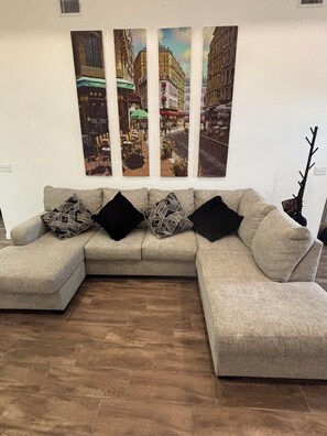 A living room with a beige sectional sofa, three black cushions, and a painting of a street scene above. - Private Room in Brandnew Home. Fullsize Bathroom & Kitchen. 5min from Airport (Harlingen)
