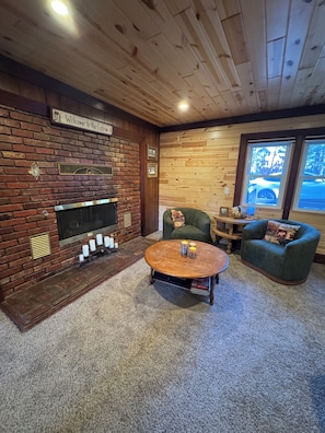 A cozy cabin-style room with a fireplace, a round coffee table, two armchairs, and a view of a parked car outside. - Family game room, great views, & steps to marina! (Big Bear Lake)