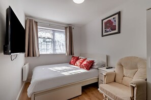 Ein Schlafzimmer mit einem Bett, einem Sessel, einem Fernseher, einem Fenster mit Vorhängen und einem gerahmten Bild an der Wand.