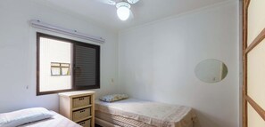 A small, sparsely furnished room with a single bed, a window with blinds, a ceiling fan, and a wall-mounted mirror. - Apartamento em Ubatuba a 50m da Praia Grande (Ubatuba)