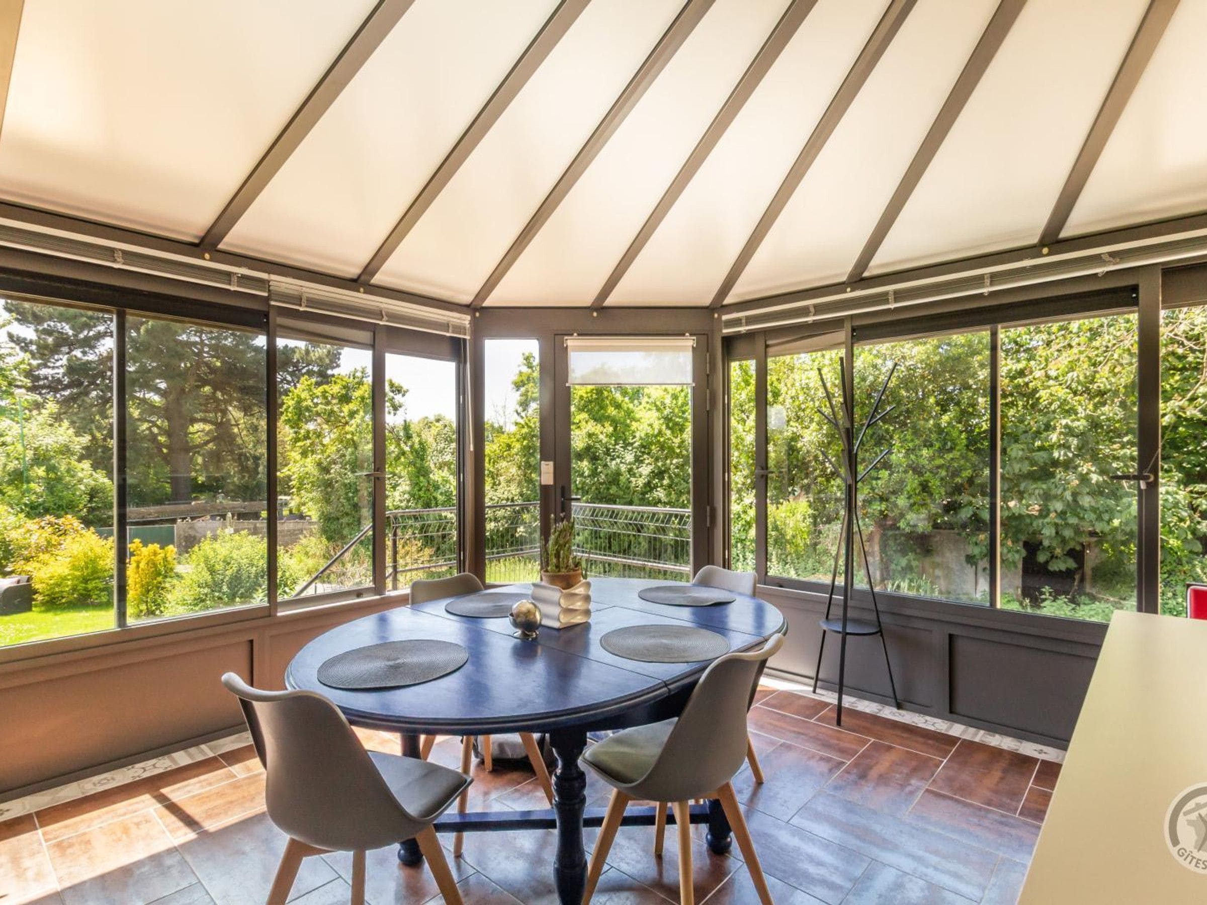 A conservatory with a round table and chairs, large windows, and a view of greenery.