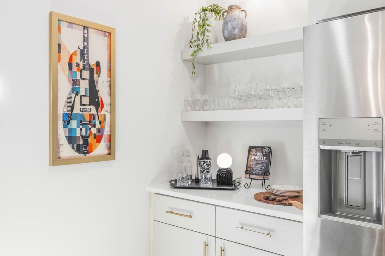A kitchen with a refrigerator, a coffee machine, a glass display, and a framed artwork.