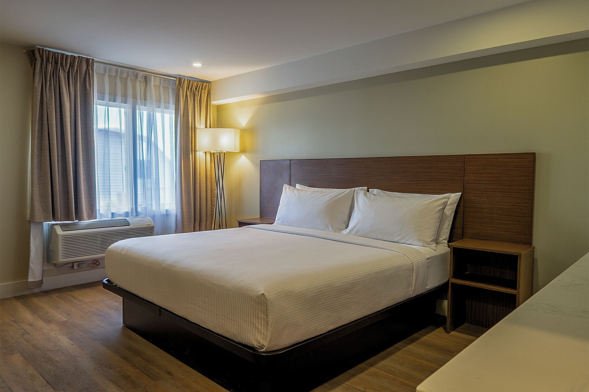 A hotel room with a bed, a nightstand, a lamp, a window with curtains, and an air conditioning unit.