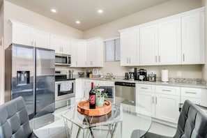 A modern kitchen with white cabinets, stainless steel appliances, and a glass dining table. - Gather & Unwind | Patio | Ramada, BBQ | 3BR (Tucson)
