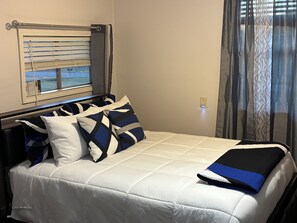 A bed with white bedding and blue and white pillows, a window with blinds, and a power outlet on the wall. - Rustic Guesthouse on the Arkansas River  White Hall. (White Hall)