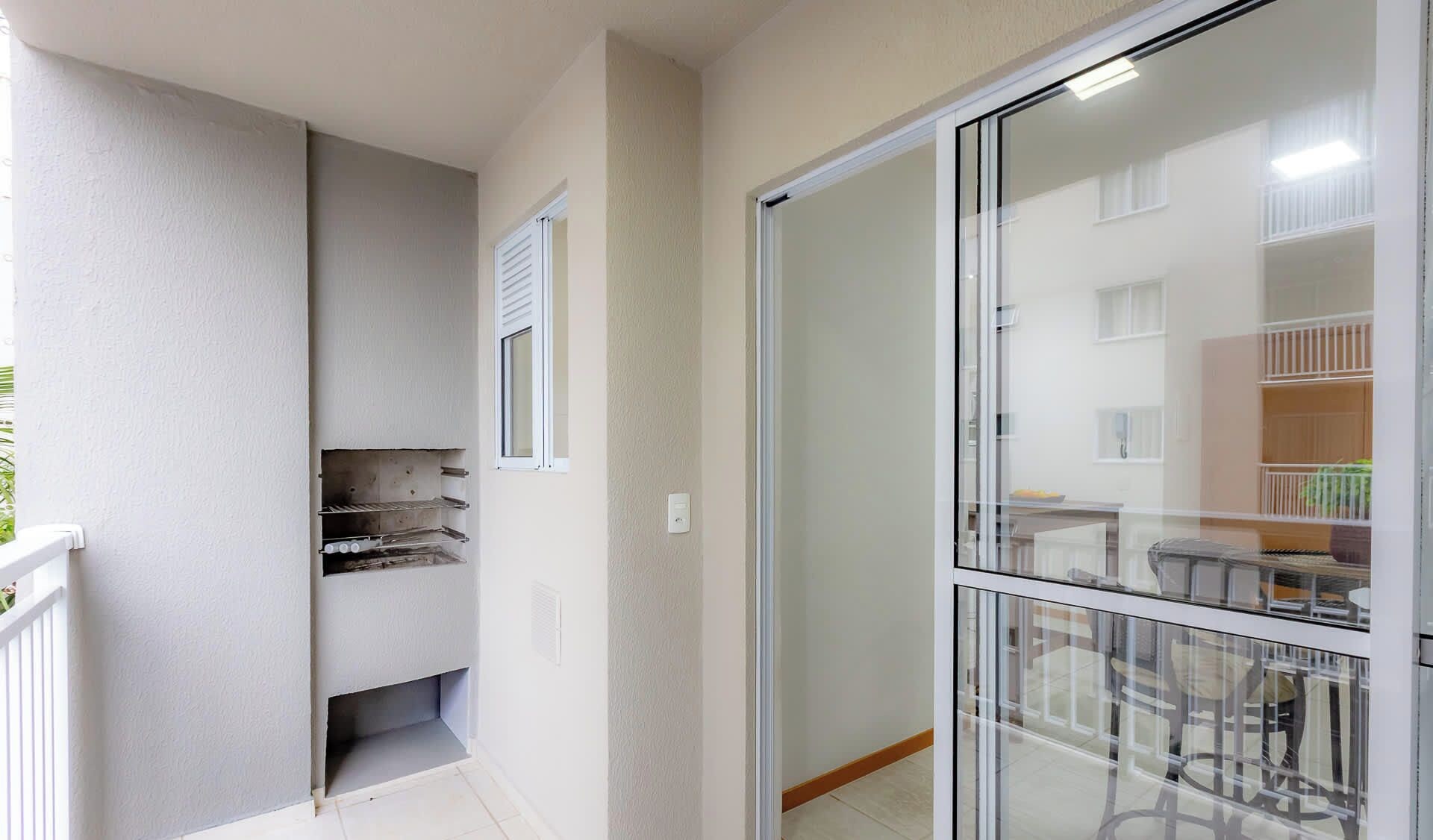 A balcony with a white railing, a small enclosed space with shelves, and a sliding glass door leading to a room with a visible kitchen area.