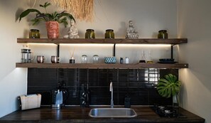 Una cocina con salpicadero oscuro, estantes de madera con botes y botellas varios, una cafetera y un lavabo.