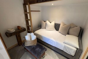A small, cozy room with a bed, a bench, a shelf with dishes, and a basket of towels. - Tiny home with gazebo (San Rafael)