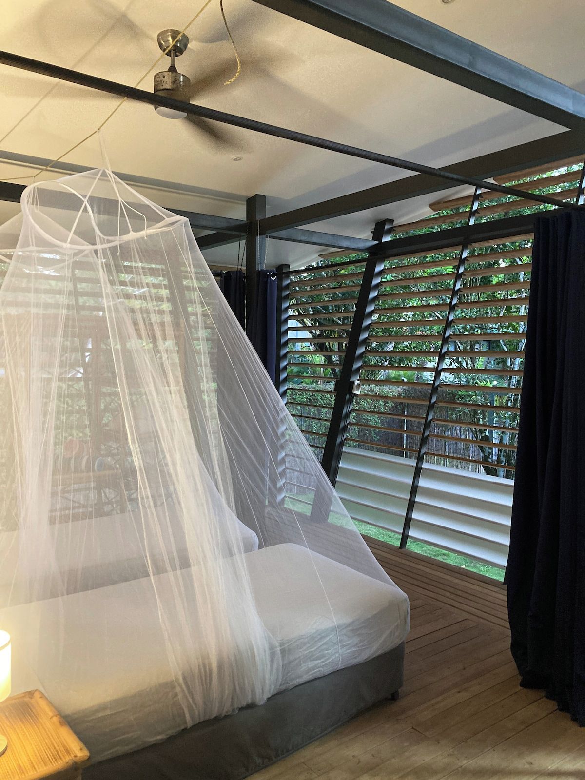Ein Schlafzimmer mit Bett, Deckenventilator, Fenster mit Jalousien und Blick ins Grüne.
