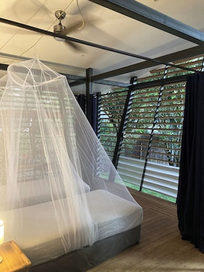 Ein Schlafzimmer mit Bett, Deckenventilator, Fenster mit Jalousien und Blick ins Grüne.