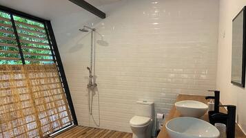A modern bathroom with a wooden floor, white tiled walls, a shower, a toilet, and two large white bowls on a wooden counter.
