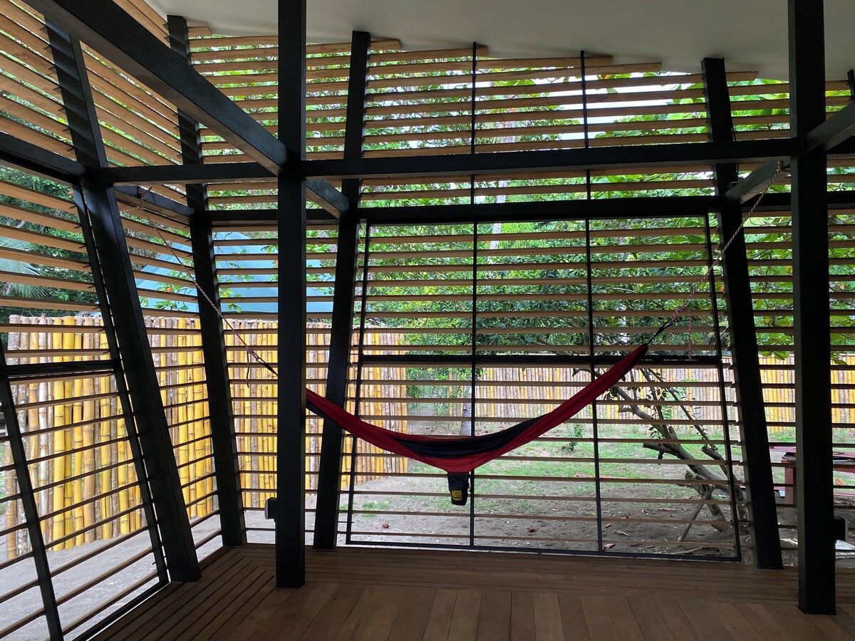 A hammock suspended between metal bars in a room with wooden flooring and a view of a bamboo fence outside.