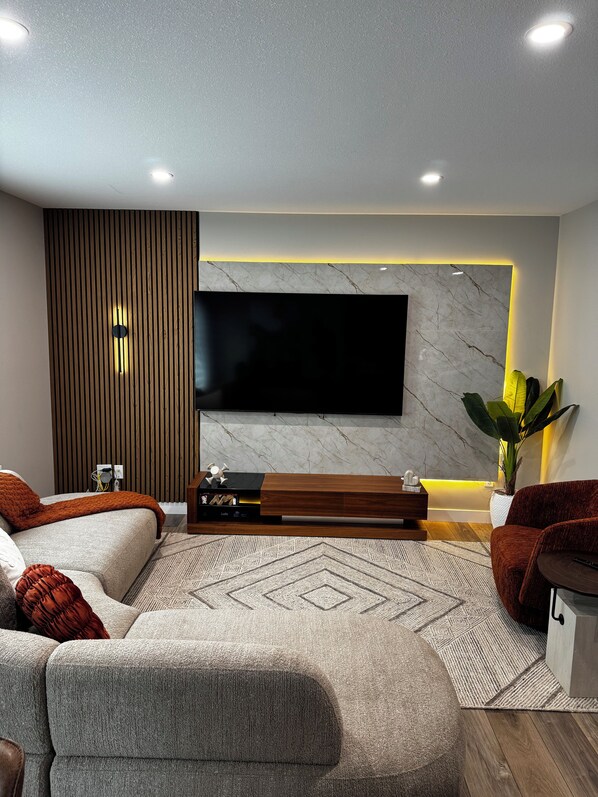 Een moderne woonkamer met een flatscreen televisie aan een marmeren muur, een houten accentmuur en een grote hoekbank.