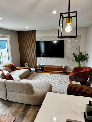 A modern living room with a sectional sofa, a flat-screen TV mounted on a marble wall, a wooden coffee table, and a dining table with a light-colored top. - Maple House | Stylish Stay for 8 (Regina)