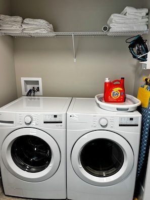 Two front-loading LG washing machines with a shelf above containing folded white towels and a red Tide detergent bottle. - Maple House | Stylish Stay for 8 (Regina)