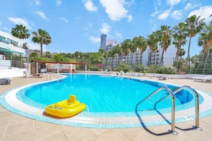 Piscina com posto de salva-vidas, cadeiras de sol e palmeiras ao fundo.