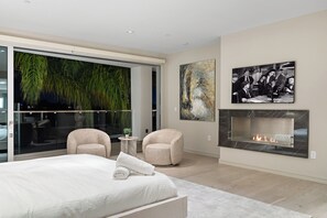 Een moderne slaapkamer met een groot bed, twee fauteuils, een open haard en een groot raam met uitzicht op groen.