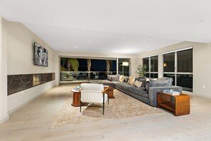 Een ruime woonkamer met een grote grijze bank, een witte fauteuil en een salontafel. Er is een groot raam met uitzicht op palmbomen en een balkon.
