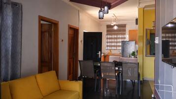 A living room with a yellow sofa, a dining area with chairs and a table, and a kitchen in the background.